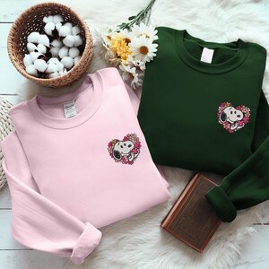 Könnte beinhalten: Zwei Sweatshirts, eines in Hellrosa und eines in Dunkelgrün, jeweils mit einem Snoopy-Motiv in einem herzförmigen Blumendesign. Sie werden auf einer weißen, fellartigen Oberfläche präsentiert, mit einem Korb mit Wattebällchen und Blumen im Hintergrund.