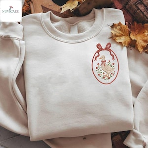 Könnte beinhalten: Cremefarbenes Sweatshirt mit einem gestickten ovalen Design. Das Design zeigt eine Ente mit roter Schleife, umgeben von roten Blumen und grünen Blättern, mit einem roten Band oben. Das Sweatshirt ist gefaltet, mit einem Markenlogo in der Ecke.