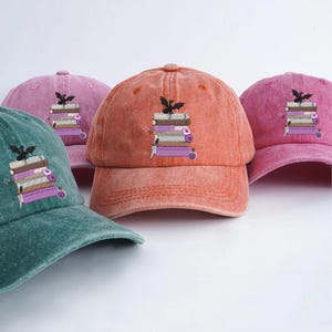 Peut inclure: Plusieurs casquettes de baseball de couleurs variées, notamment turquoise, rose et orange. Chaque casquette présente un motif brodé d'une pile de livres surmontée d'une chauve-souris. Les casquettes sont en tissu doux et texturé.