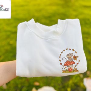 Può includere: Felpa bianca con una grafica circolare di un topo dei cartoni animati seduto su un fungo rosso e bianco, che tiene foglie autunnali. Il disegno è incorniciato da una ghirlanda di foglie. La felpa ha uno scollo tondo.