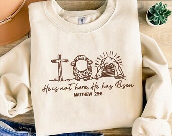 May include: Cream-colored sweatshirt with a brown graphic of a cross, lamb, and tomb. The text "He is not here, He has Risen" and "MATTHEW 28:6" are printed below the graphic. The sweatshirt is displayed on a wooden surface.