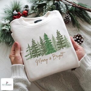 Könnte beinhalten: Cremefarbenes Sweatshirt mit einem Wald aus grünen Kiefern und dem Schriftzug "Merry & Bright" in goldener Schrift. Das Sweatshirt ist gefaltet und wird in Händen gehalten, mit Weihnachtsgrün im Hintergrund.
