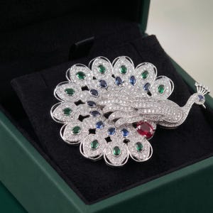 Peut inclure: Broche en forme de paon ornée de pierres précieuses, présentée dans un écrin vert. Le paon est orné de pierres claires, vertes, bleues et rouges. Les plumes de la queue du paon sont déployées, révélant un travail de joaillerie détaillé.