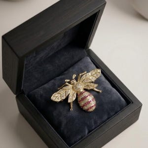 May include: A bee-shaped brooch crafted from gold-toned metal, featuring wings and a head encrusted with clear, sparkling stones. The abdomen displays alternating horizontal bands of clear stones and deep pink, rectangular-cut stones.