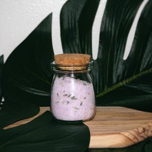 Peut inclure: Un petit pot en verre transparent rempli de sels de bain de couleur lavande et surmonté d'un bouchon en liège. Le pot est posé sur une surface en bois, avec de grandes feuilles vertes en arrière-plan. Les sels de bain contiennent de petits bourgeons de lavande visibles.