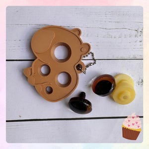 May include: A brown bear-shaped keychain with multiple circular cutouts, a small chain, and several craft supplies, including two brown and one yellow plastic eye. The items are arranged on a white wooden surface.