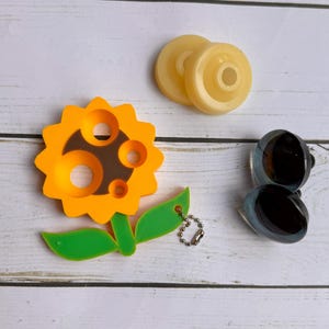 May include: An assortment of decorative items on a white wooden surface. The items include a yellow sunflower-shaped keychain, two yellow circular objects, and a pair of dark blue and black oval-shaped objects. The sunflower has green leaves.