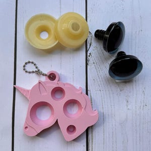 May include: An assortment of craft supplies including two yellow plastic discs, two black plastic safety eyes, and a pink unicorn-shaped keychain. The items are arranged on a white wooden surface.