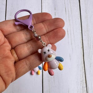 May include: A whimsical keychain featuring a small, pink teddy bear charm with colorful arms and legs. The bear has black cross-shaped eyes and a brown nose. It is attached to a purple clasp with a beaded chain.
