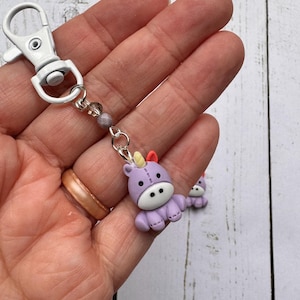 May include: A purple unicorn charm with a white belly, black eyes, and a colorful horn is attached to a silver chain and a white metal clasp. The charm is held in a hand.