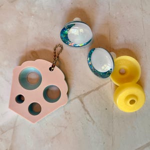 May include: A collection of craft supplies including two pairs of iridescent earrings, a pink and blue stencil, and two yellow spools. The earrings have a white top and a blue and green glitter base. The stencil is pink with several circular cutouts.