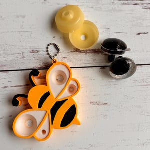 May include: A yellow and black bee-shaped keychain with a silver chain, along with two yellow plastic pieces and two black plastic eye pieces. The bee has white and orange details. The items are on a light wood surface.