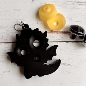 May include: A black dragon-shaped keychain with a silver chain, featuring circular openings for attaching accessories. Two yellow plastic pieces and two black plastic eyes are also present, suggesting a crafting or DIY theme.
