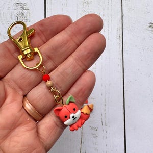 May include: A red fox keychain with a gold-colored clasp and chain. The fox charm has white and black accents, with green ears and a yellow and orange tail. The keychain also features red and tan beads.