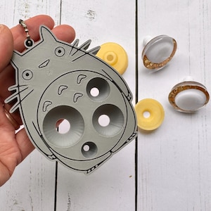 May include: A gray Totoro-shaped keychain with a black outline, held against a white wooden surface. Two yellow donut-shaped objects and two white and gold-rimmed objects are also present. The keychain has a silver clasp.