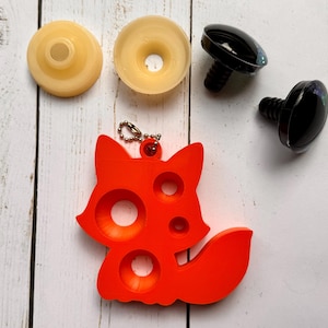 May include: An assortment of craft supplies including two light yellow plastic pieces, two black plastic safety eyes, and a bright orange fox-shaped charm with three circular cutouts. The items are arranged on a white wooden surface.