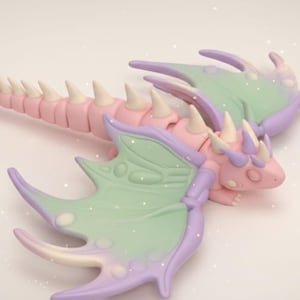 Könnte beinhalten: Pastellfarbene Drachenfigur mit rosa Körper, weißen Stacheln und lila Flügeln. Die Flügel sind eine Mischung aus Grün und Lila. Der Drache liegt ausgestreckt, mit ausgestrecktem Schwanz. Das Design ist skurril und fantasy-orientiert.