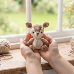 May include: A small, crocheted fawn figurine featuring a light tan and cream-colored yarn body with pointed ears, dark eyes, and a stitched nose. The toy is accompanied by a tan yarn ball, a wooden crochet hook, and a tag labeled HANDMADE CROCHET ANIMAL PATTERN - INSTANT DOWNLOAD.