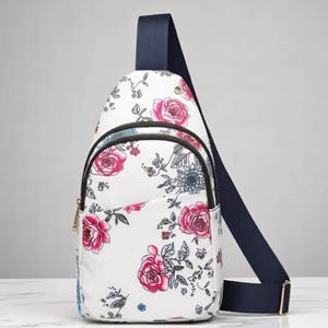 Può includere: Borsa a tracolla bianca con un motivo floreale di rose rosa e foglie verdi. La borsa ha una tracolla blu navy e una cerniera dorata. La borsa è progettata per essere indossata a tracolla.