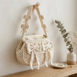 Può includere: Una borsa all'uncinetto color crema con chiusura a scatto in metallo, un manico strutturato decorato con piccoli fiocchi e una patta frontale ornata da due grandi fiocchi con lunghi nastri pendenti, il tutto realizzato in filato testurizzato.