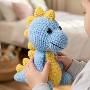 Puede incluir: Un juguete de dinosaurio de ganchillo azul y amarillo con grandes ojos negros y una boca sonriente. El peluche tiene púas amarillas en la espalda y lo sostiene una persona.