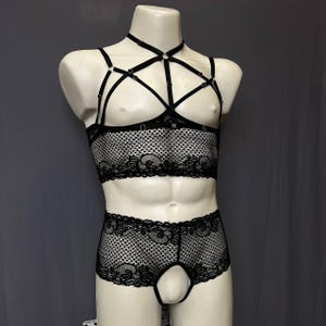 May include: A black lace lingerie set consisting of a cropped top and matching shorts. The top features a complex system of adjustable straps forming a harness design over the chest. Both pieces are made of a sheer, patterned lace fabric.