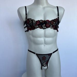 May include: Black lingerie set featuring floral embroidery in red, brown, and white. The set includes a bra with adjustable straps and a matching thong. The bra has a harness-style design.