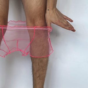 May include: A pair of sheer, translucent pink underwear featuring a delicate, ruffled lettuce-edge trim along the waistband and leg openings. The garment is constructed from a lightweight, fine mesh fabric with visible neon pink stitching.
