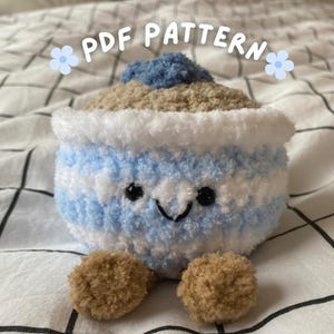 Puede incluir: Un juguete de peluche con forma de cupcake sonriente con un cuerpo a rayas azules y blancas, una parte superior beige y un pompón azul. El juguete tiene ojos negros y una sonrisa cosida. El texto "PDF PATTERN" está encima del juguete.