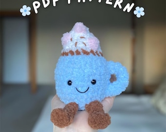 Könnte beinhalten: Eine hellblaue Plüsch-Kaffeetasse mit einem lächelnden Gesicht, braunen Füßen und einem Schlagsahne-Topping mit rosa Akzenten. Der Text "PDF PATTERN" befindet sich über der Tasse, mit blauen Blumenakzenten.