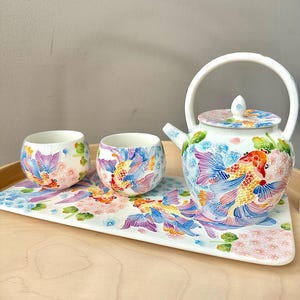 Può includere: Un servizio da tè in ceramica con una teiera, due tazze e un vassoio rettangolare. Il set è decorato con motivi di pesci koi e floreali nei toni del blu, rosa e arancione. La teiera ha un manico curvo. Il servizio da tè è su un vassoio di legno.