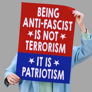 Puede incluir: Una persona sostiene un cartel rojo y azul con texto blanco: "SER ANTIFASCISTA NO ES TERRORISMO ES PATRIOTISMO". El cartel tiene una parte superior roja y una inferior azul, con estrellas blancas.