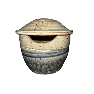 May include: A ceramic container with a lid. The container is beige with blue stripes and speckled detailing. The lid has a rounded shape with horizontal ridges. The container has a small opening on the side.