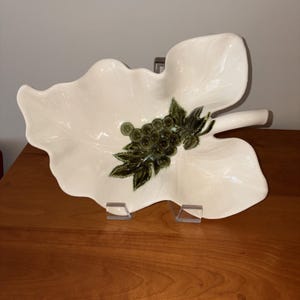 May include: A cream-coloured ceramic dish shaped like a large leaf with a textured surface. The centre of the dish features a raised, dark green relief design of a cluster of grapes with accompanying leaves, adding a decorative element to the piece.