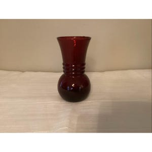 May include: A dark red glass vase with a bulbous base and a flared top. The vase has a series of horizontal ridges around the middle. The vase is sitting on a white surface.