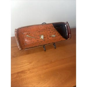 Puede incluir: Bandeja rectangular de cerámica con un esmalte moteado naranja y marrón. La bandeja presenta una esquina negra y un diseño decorativo de pájaros y hojas. La bandeja está soportada por un soporte de metal negro.