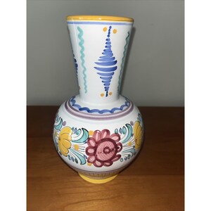 May include: A white ceramic vase with a rounded body and a flared neck. The surface features hand-painted floral motifs in yellow, red, and teal, accented by blue vertical patterns on the neck and a yellow rim at the top and base of the vessel.