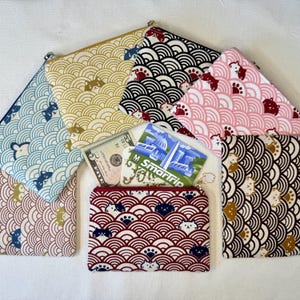 May include: A collection of zippered pouches in various colors, featuring a repeating wave pattern with cat and paw prints. The pouches are in shades of blue, yellow, pink, black, and red. A five-dollar bill and a transit card are visible.