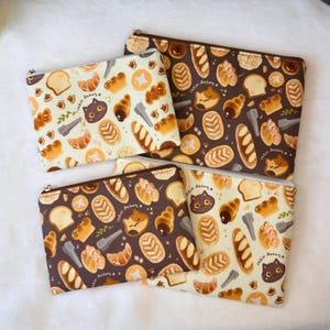 May include: Four zippered pouches with a repeating pattern of baked goods and a cat face. The pouches are in two color schemes: cream and brown. The text "Cat Bakery" is visible on the pouches.