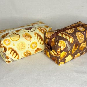 May include: Two rectangular zippered pouches with a bakery theme. One is light yellow with various bread illustrations, the other is brown with similar bread designs. Both have brown zippers and a small charm.