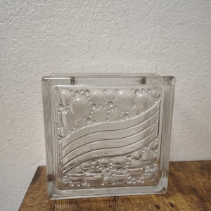 May include: A clear glass block with a decorative wave and bubble design. The block is square and sits on a wooden surface. The glass has a textured appearance.