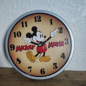 May include: Round wall clock featuring Mickey Mouse. The clock face has a tan background with black numbers and the words "Mickey Mouse" in red. Mickey Mouse is in the center, pointing with his fingers. The clock has a silver rim.