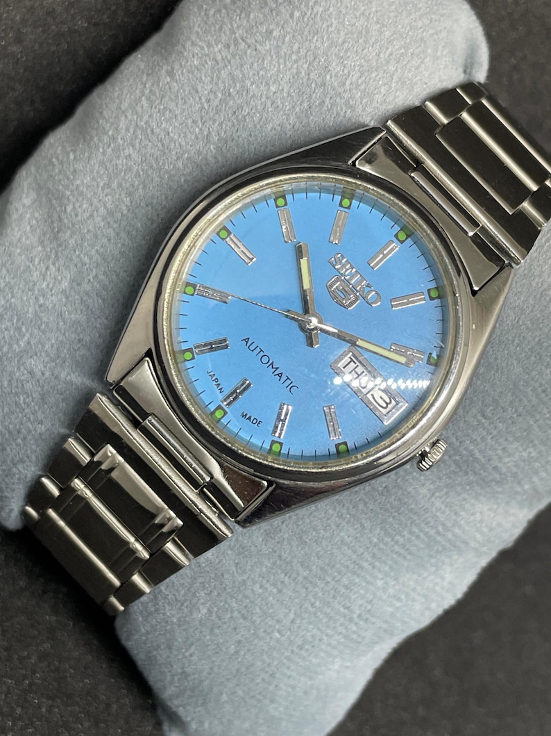 Pu&ograve; includere: Un orologio automatico Seiko 5 in acciaio inossidabile con quadrante azzurro. Il quadrante mostra il giorno e la data, con dettagli verdi e argento. Il cinturino dell'orologio &egrave; in acciaio inossidabile.