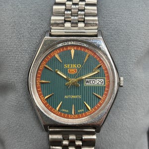May include: A vintage Seiko 5 automatic watch with a silver-tone metal band and case. The watch face is teal with gold-tone hands and markers, and a red outer ring. The day and date are displayed at the 3 o'clock position.