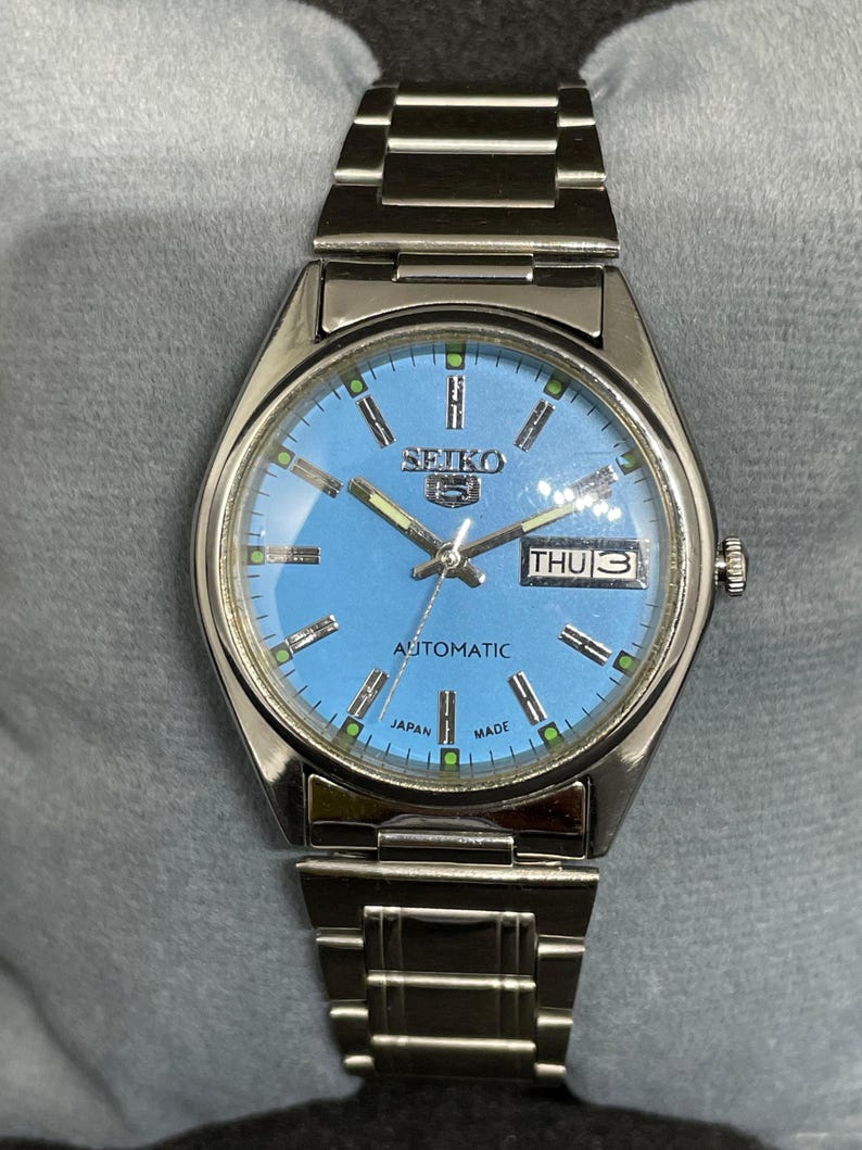 Pu&ograve; includere: Un orologio da polso automatico Seiko color argento con quadrante azzurro. L'orologio presenta un display giorno-data e un cinturino a maglie argentate. Il quadrante ha dettagli verdi e argento, con il testo "SEIKO" e "AUTOMATIC".