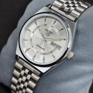 May include: A silver Seiko 5 automatic wristwatch with a silver dial and a stainless steel band. The watch face displays the day and date, with the day reading "SAT 25". The watch has a silver bezel and a crown on the right side.