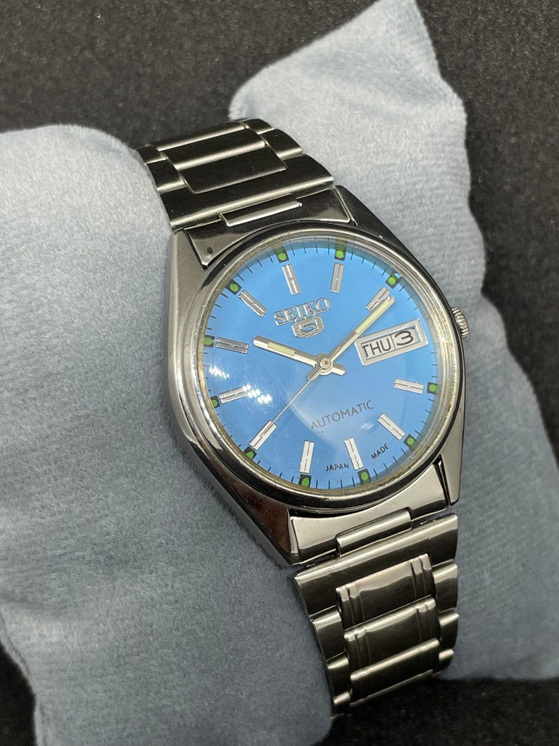 Pu&ograve; includere: Un orologio automatico Seiko 5 in acciaio inossidabile con quadrante azzurro. Il quadrante mostra il giorno e la data, con dettagli verdi e argento. Il cinturino dell'orologio &egrave; in acciaio inossidabile.