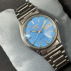 Pu&ograve; includere: Un orologio automatico Seiko 5 in acciaio inossidabile con quadrante azzurro. L'orologio presenta un display giorno e data, lancette e indici luminescenti e un bracciale color argento. Il quadrante dell'orologio riporta la scritta "Seiko 5 Automatic Japan Made".