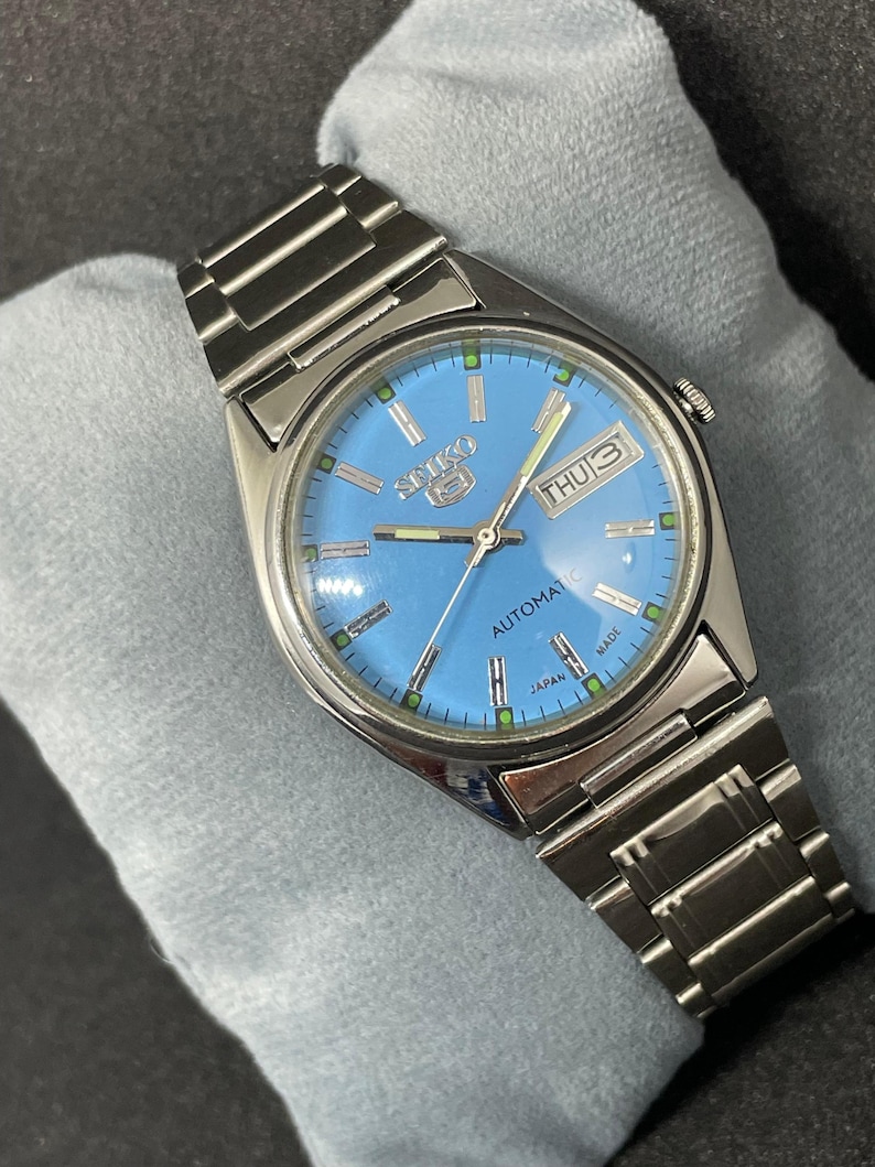 Pu&ograve; includere: Un orologio automatico Seiko 5 in acciaio inossidabile con quadrante azzurro. L'orologio presenta un display giorno e data, lancette e indici luminescenti e un bracciale color argento. Il quadrante dell'orologio riporta la scritta "Seiko 5 Automatic Japan Made".