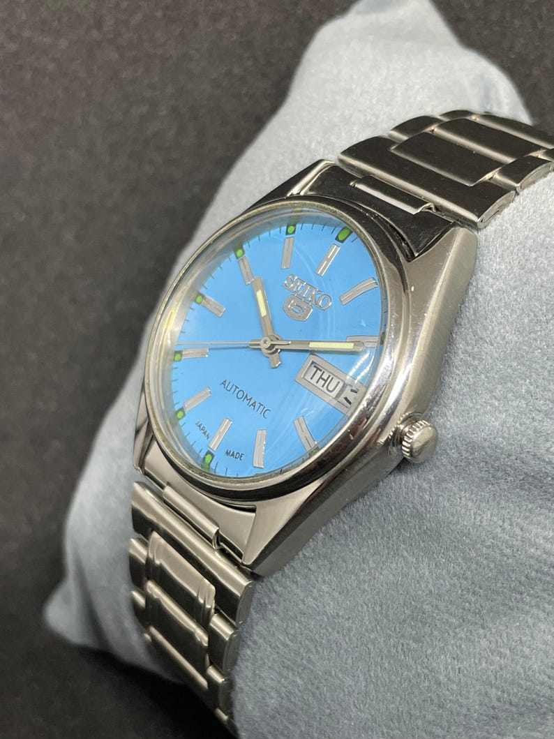 Pu&ograve; includere: Un orologio automatico Seiko 5 argentato con quadrante azzurro. L'orologio presenta un cinturino argentato e un display giorno-data. Il quadrante ha lancette e indici argentati. L'orologio &egrave; contrassegnato "AUTOMATIC" e "JAPAN MADE".